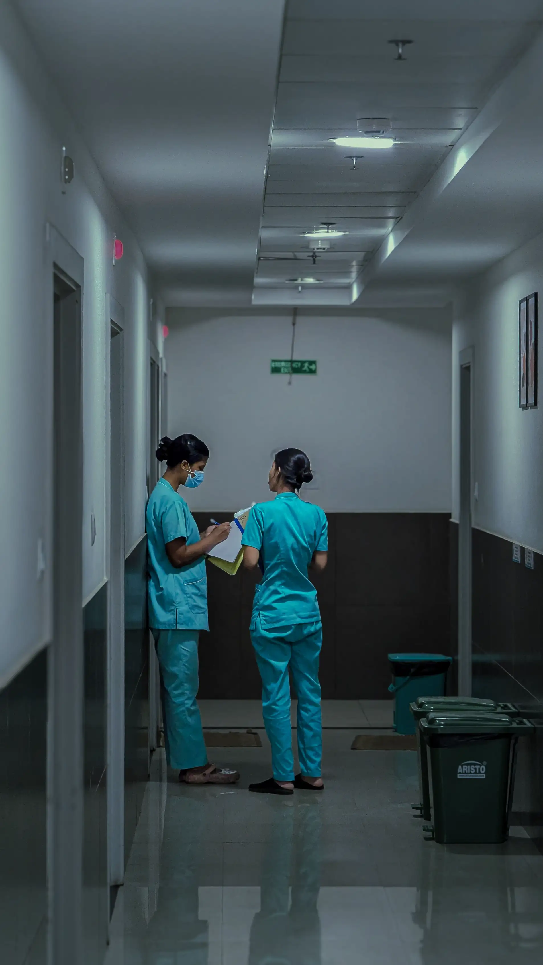 Two nurses in teal scrubs reviewing a patient chart in a dim hospital hallway.